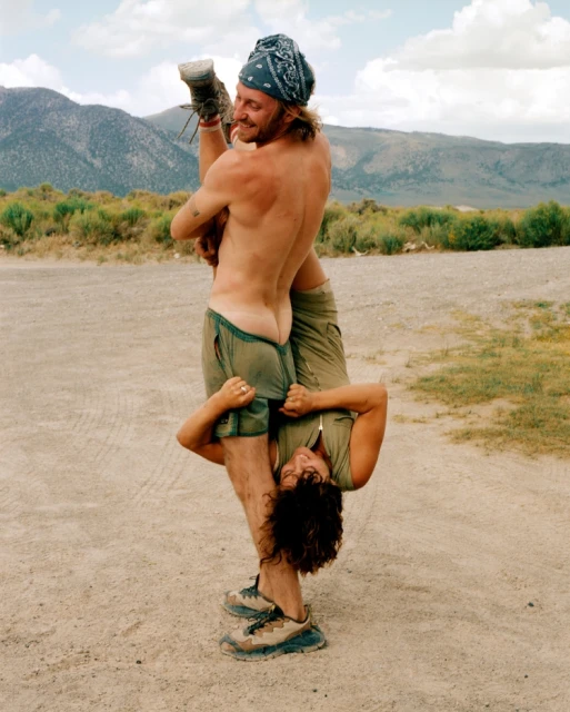 Man holding woman upside down in rural landscape. He's shirtless with shorts and a bandana; she wears a zip-up shirt and shorts.