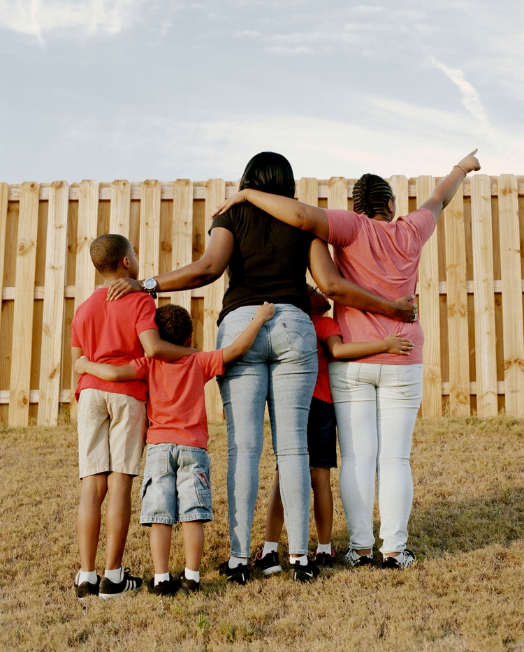 Family of five with two adult women and three children embrace while facing away from the camera in a yard with a wood fence.