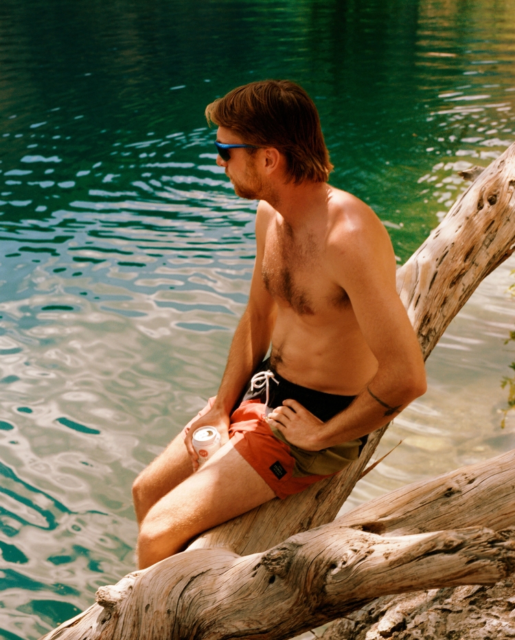 Man sitting shirtless on a fallen tree trunk over turquoise water holding a beverage can, by Peyton Fulford