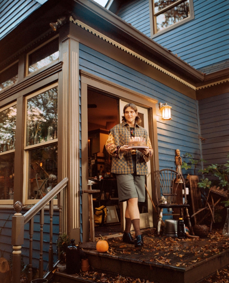 Adult female stands on a porch holding a cake with candles, surrounded by plants, furniture, and a blue clapboard house.