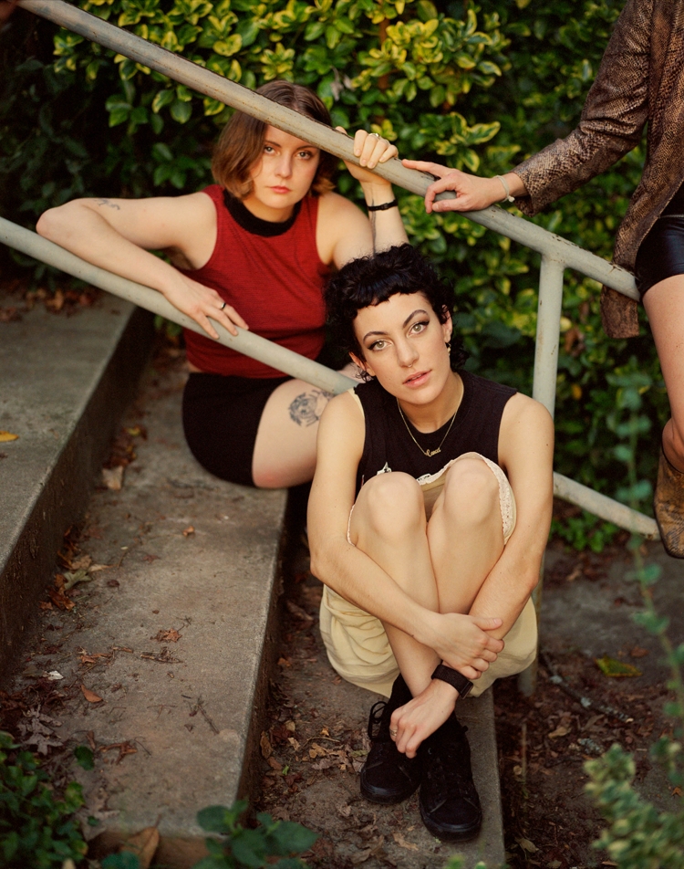 Two young women sit on concrete steps with a handrail in a green outdoor setting, posing for a portrait in casual clothing.