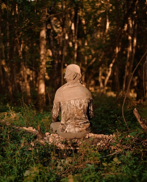 Person in camouflage clothing sits on a log in a dense forest, back to camera, surrounded by lush green vegetation and trees.