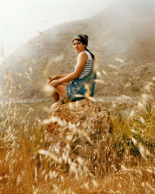 Young woman in denim shorts and bandana sitting on a rock in a grassy field with mountains in the background on a hazy day.