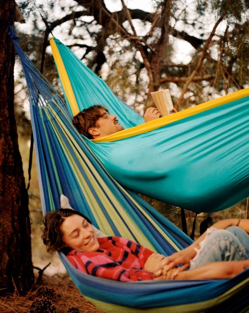 Two people relaxing in separate hammocks in a wooded area, one reading a book and the other smiling with interlaced fingers, daytime.
