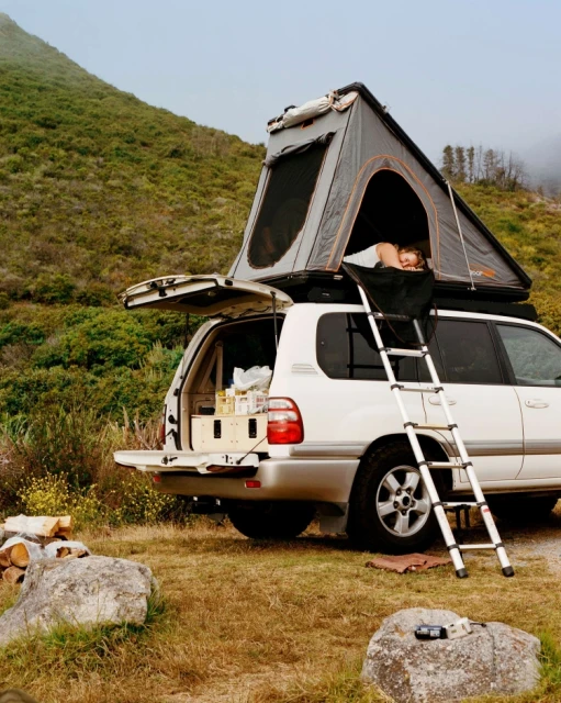 SUV parked in a grassy field with a roof tent, with one adult inside the tent, ready for camping in a rural setting near a grassy mountain.