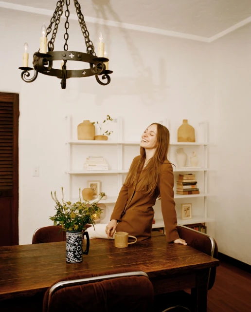 A woman in a brown blazer sits on a wood table with flowers and a mug, illuminated by a chandelier in an indoor setting with shelves.