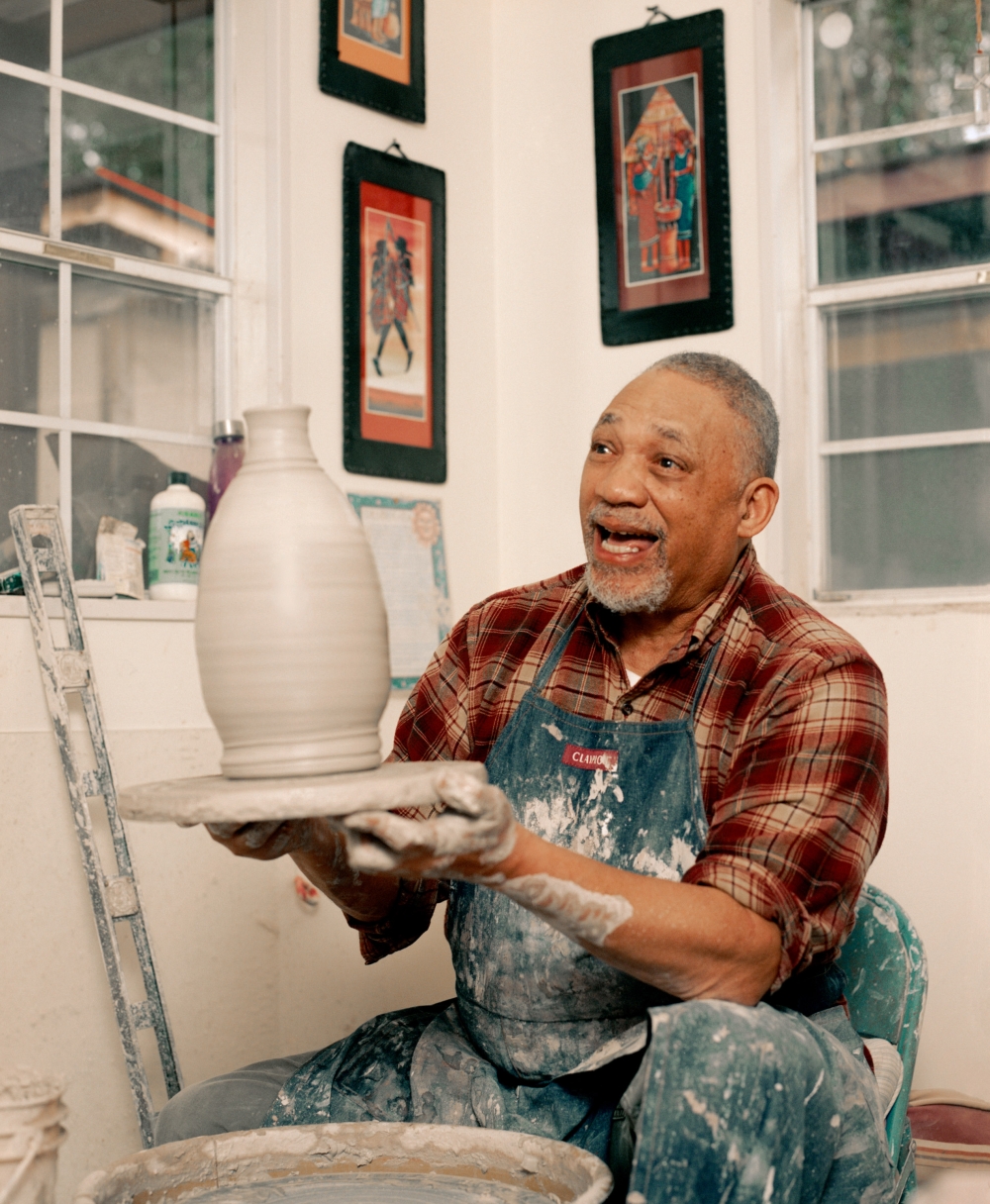 An elderly African-American man is shown in his pottery studio, holding up a vase he made on the pottery wheel. Indoor shot.