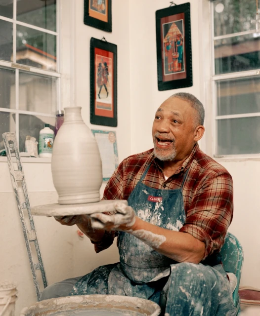An elderly African-American man is shown in his pottery studio, holding up a vase he made on the pottery wheel. Indoor shot.