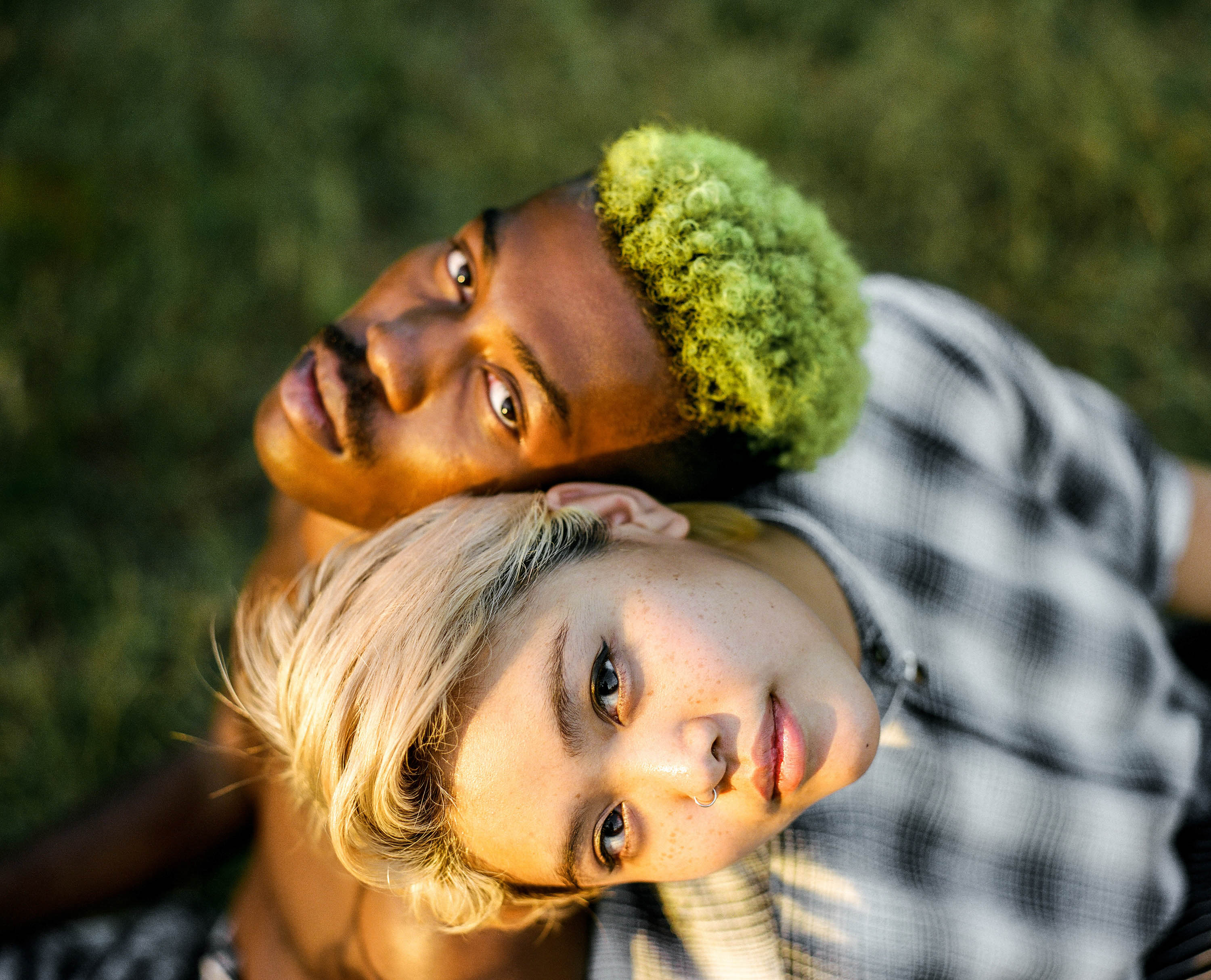 Eye-level shot of two people, one with dyed green hair, lying side-by-side in an outdoor grassy area, looking up toward the camera.