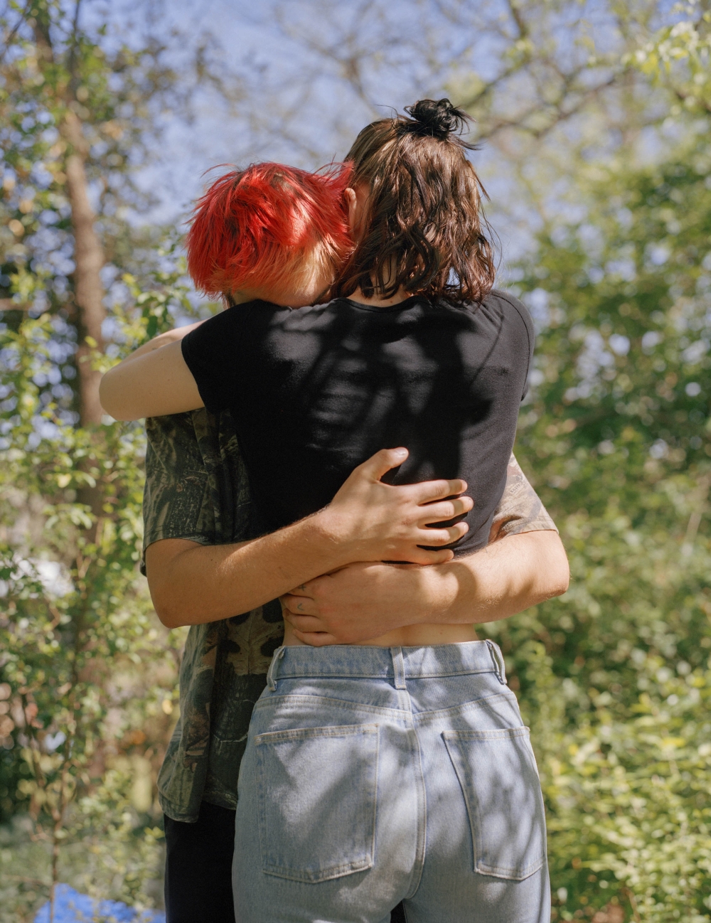 Two people embrace in a sunlit outdoor setting; one with short red hair, the other with dark hair in a bun, surrounded by lush green foliage.