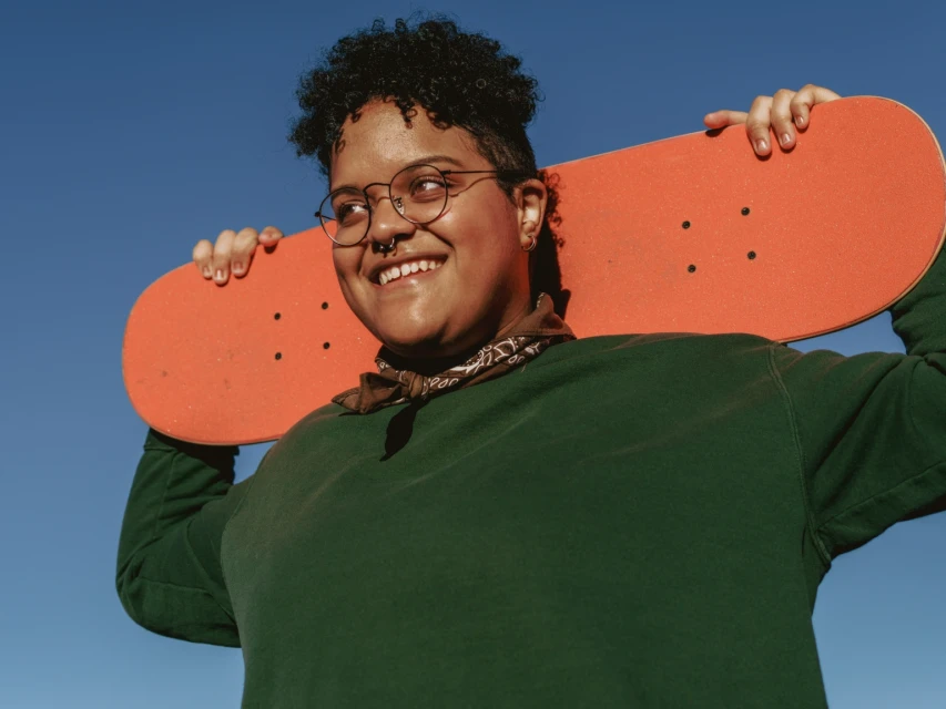Smiling adult female skateboarder wearing eyeglasses and a green sweatshirt with bright blue sky in the background on a sunny day.