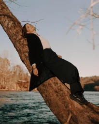 Woman in a black coat leaning back against a thick leaning tree trunk over a river during sunset.