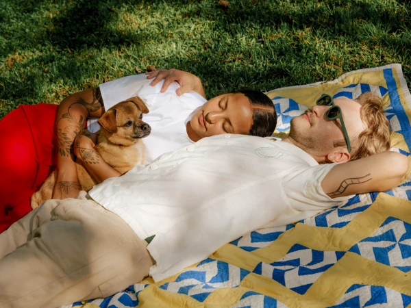 Two people lying on a yellow and blue patterned blanket on grass with a small brown dog resting between them.