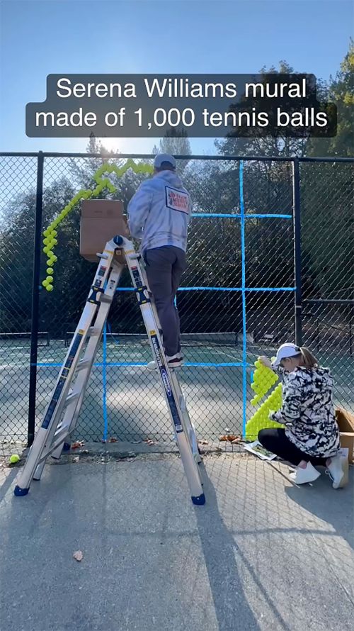 Two people assemble a Serena Williams mural made of tennis balls on a chain-link fence outside, using a ladder and a box of balls.