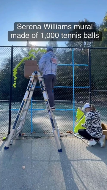 Two people assemble a Serena Williams mural made of tennis balls on a chain-link fence outside, using a ladder and a box of balls.