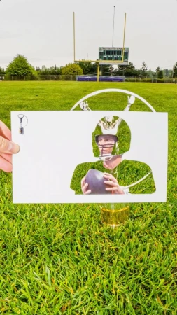 Conceptual outdoor shot of a football quarterback cut-out in a white paper over lush green grass with a football field in the background.