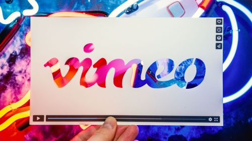 Vimeo brand logo is displayed on a white card held in hand against colorful neon lights, creating a vibrant and modern presentation.