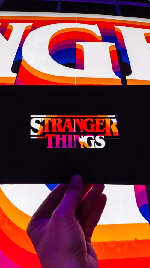 Close-up of a hand holding a 'Stranger Things' advertisement with layered rainbow-colored font and a black background.