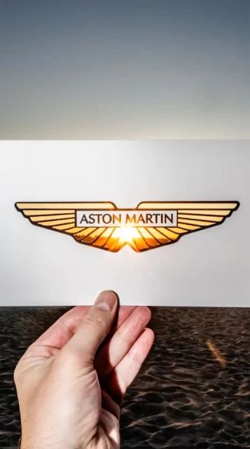 Aston Martin logo being held against a background of water and sky. Automotive design displayed by a hand over the natural setting.