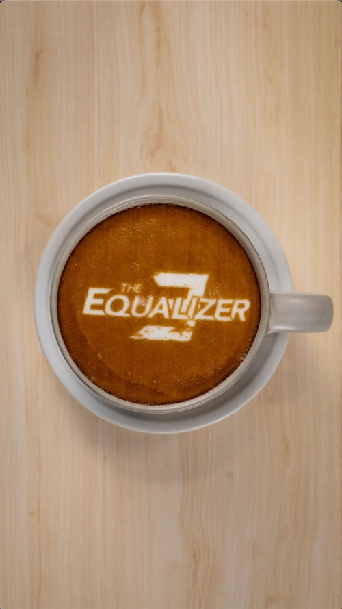 Overhead shot of a latte with white foam art depicting "The Equalizer 3" logo, sitting on a white saucer on a wood surface.