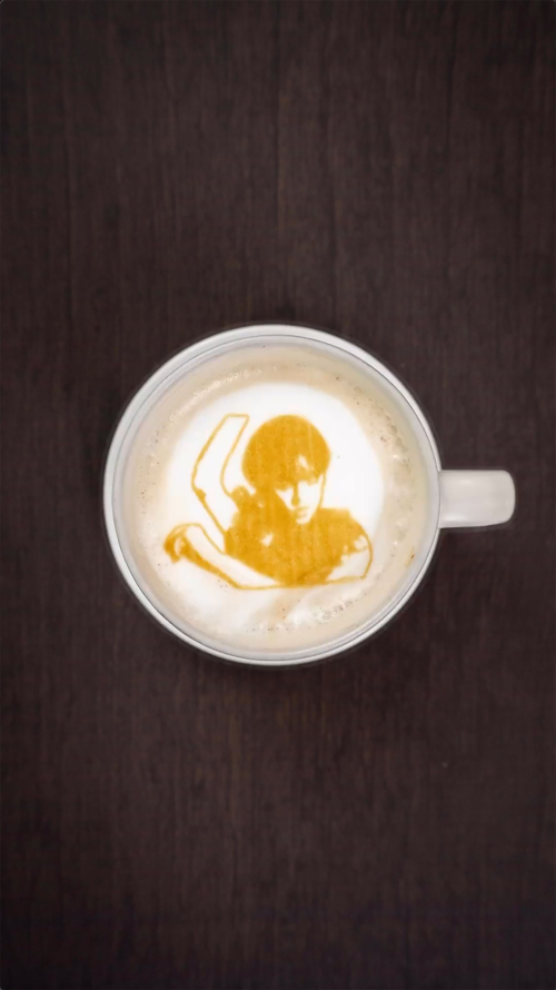 Overhead shot of a coffee cup with latte art depicting a person with their arm raised, set on a dark brown wooden surface.