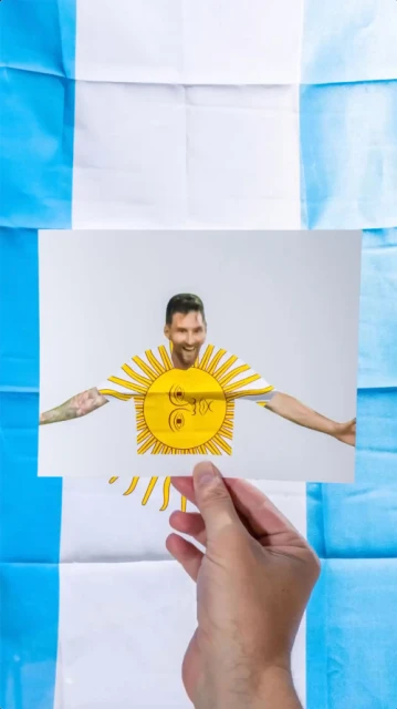 Hand holding a photo of a smiling man with a sun graphic on his t-shirt, against a light-blue and white striped flag backdrop.