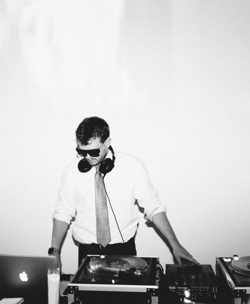 Black and white shot of a DJ in sunglasses and headphones, standing at a mixer with turntables, laptop, and white background.