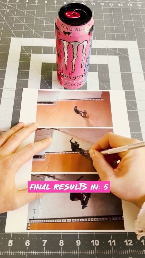 Pink Monster energy drink stands next to a print of action photos, a craft knife, and a cutting mat on a tabletop for a craft project.