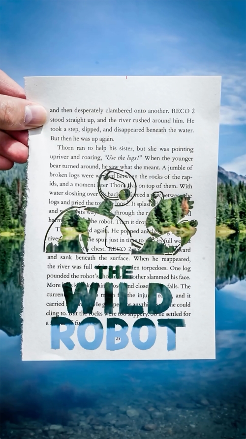 Hand holding 'The Wild Robot' book page against a backdrop of a still lake and mountain with evergreen trees on a sunny day.