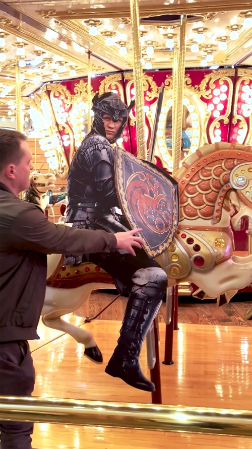 Man places hands near cutout of figure in black armor on a carousel horse under ornate lighting and a gold railing.