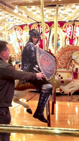 Man places hands near cutout of figure in black armor on a carousel horse under ornate lighting and a gold railing.