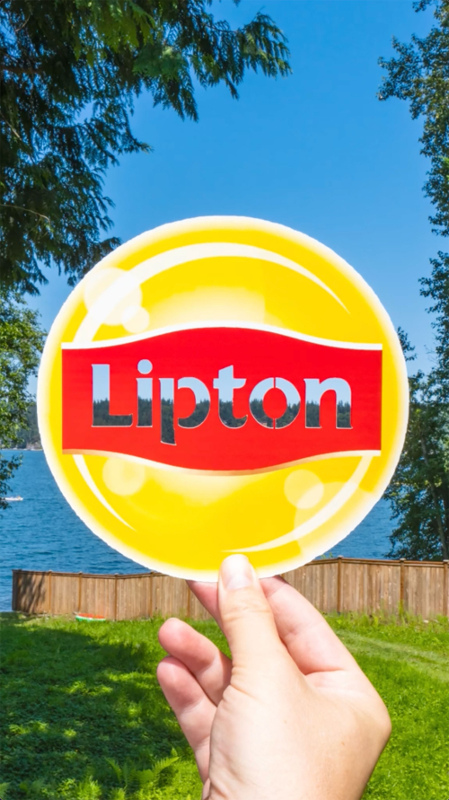 Hand holding a Lipton Iced Tea advertising sign outdoors against a bright blue sky and green trees and lawn. The sign is yellow and red.