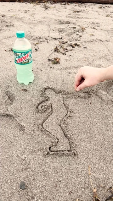 Person draws a stick figure in the sand near a bottle of Mountain Dew on a beach in daylight with an arm visible.