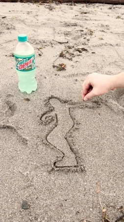 Person draws a stick figure in the sand near a bottle of Mountain Dew on a beach in daylight with an arm visible.