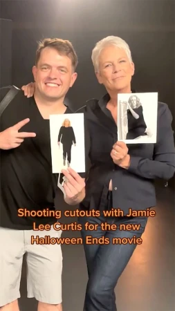 Two smiling adults, Jamie Lee Curtis, holding cutouts in a photo studio with a dark backdrop, creating Halloween Ends movie promotional content.