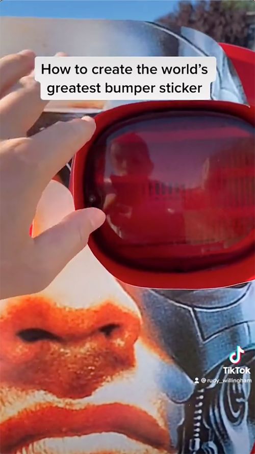 Close-up of a person holding up a large bumper sticker featuring a human face and red visor, set against a blue sky backdrop.
