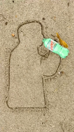 Outline of a person drawn in sand next to a partially empty bottle of Mountain Dew, with a yellow spoon on a sandy surface.