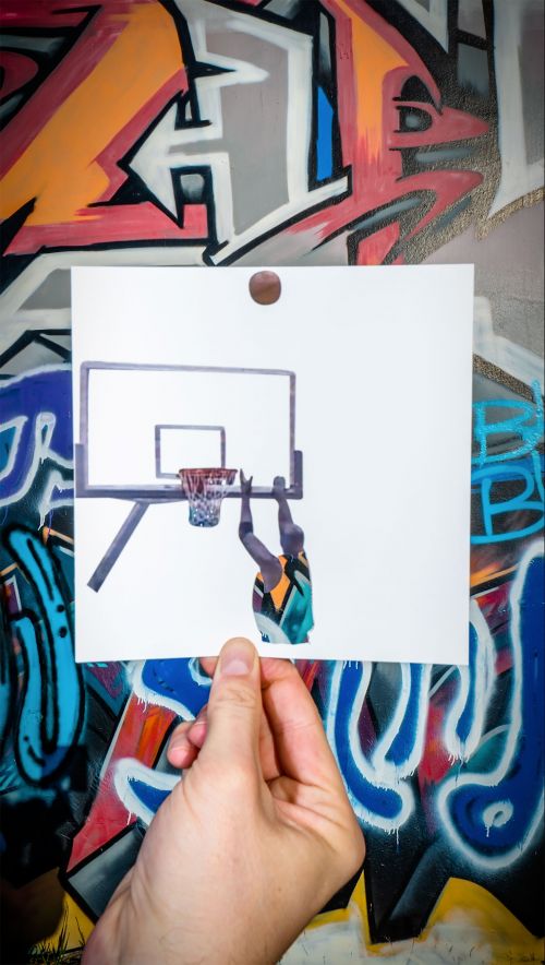A photograph of a hand holding a paper with a drawing of a basketball player dunking the ball, set against colorful graffiti art.