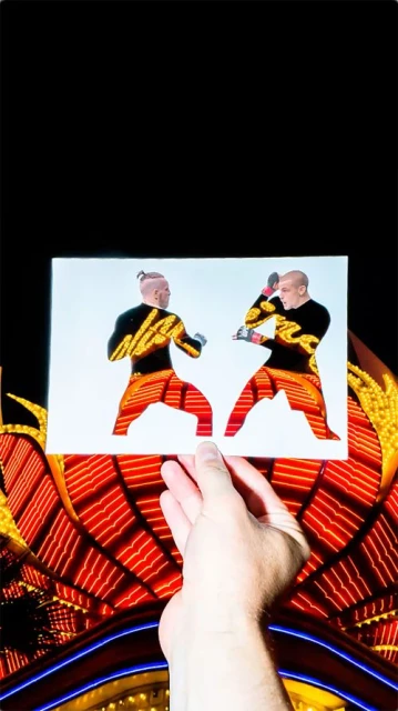 Hand holds a photograph of two male fighters with distinctive black and orange attire, set against a vibrant red and yellow light display.