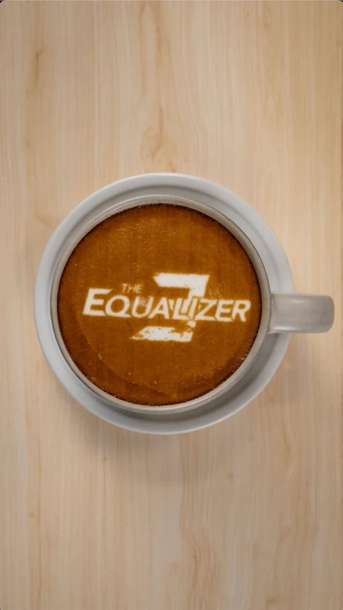 Overhead view of a white coffee mug filled with espresso and foam showing the logo for The Equalizer 3 on a light wood surface