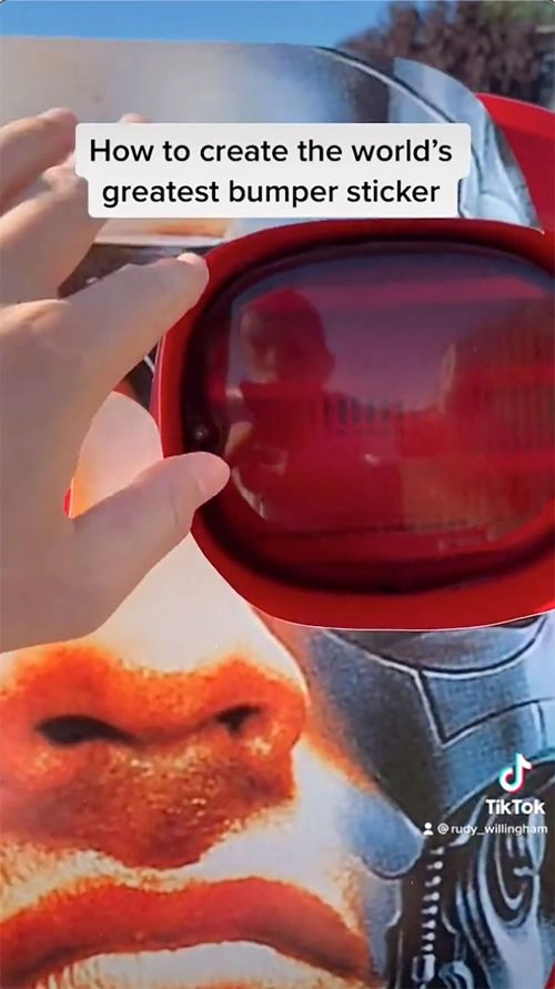 Close-up of a person hand interacting with a red car taillight and bumper sticker graphic by Rudy Willingham.