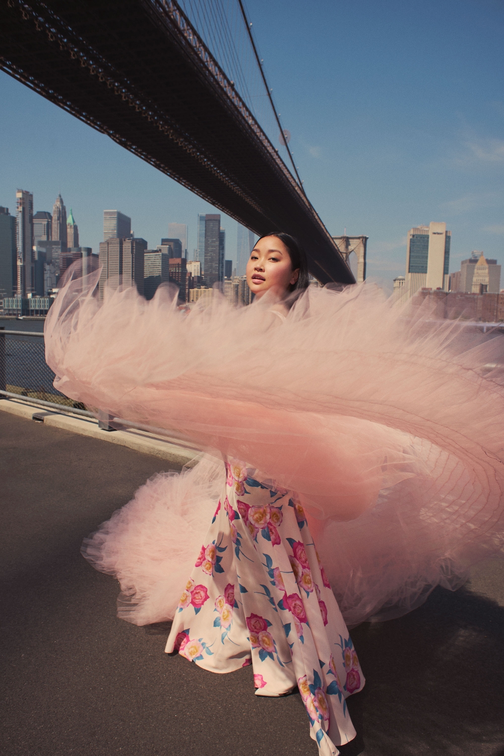 A woman in a floral skirt and pink tulle stands on a bridge with a city skyline visible in the background on a sunny day.