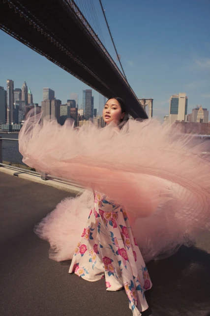 A woman in a floral skirt and pink tulle stands on a bridge with a city skyline visible in the background on a sunny day.
