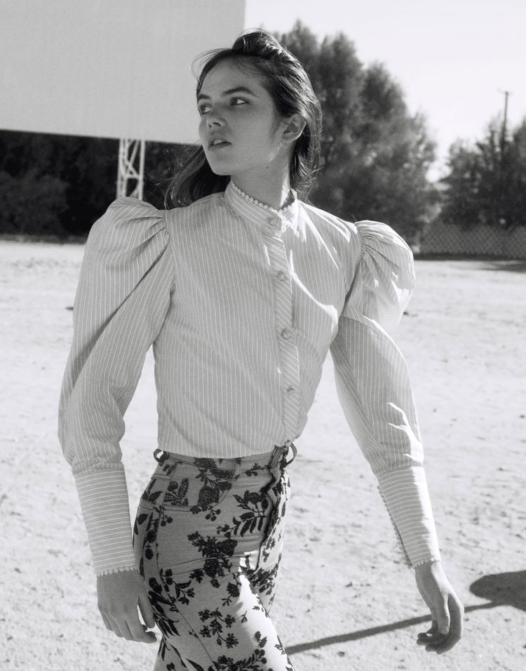 Black and white outdoor shot of a young adult woman in a striped shirt and floral pants looking away from camera.
