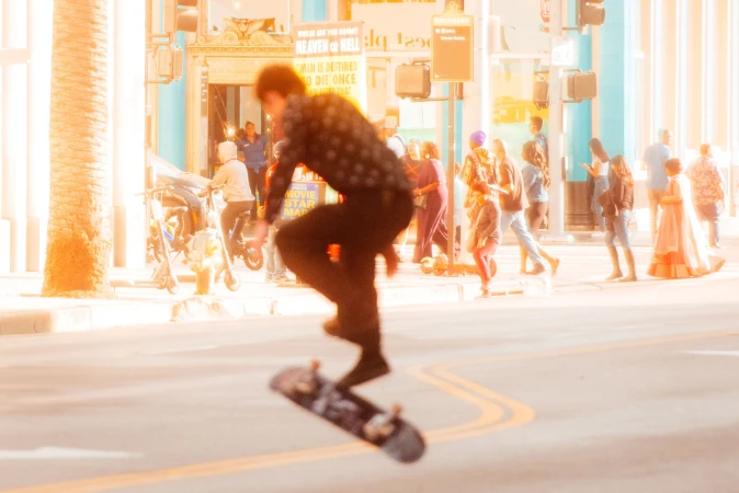 Motion-blurred skateboarder performing an aerial trick on a sunlit Los Angeles street with pedestrians in the background