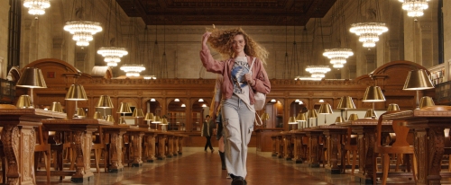 A young woman with curly blonde hair walks confidently through a grand library hall with rows of tables, lamps, and ornate architecture.