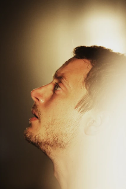 Profile of adult male with a light beard looking up in a soft-lit, dark environment with a bright glow around the head and shoulders.