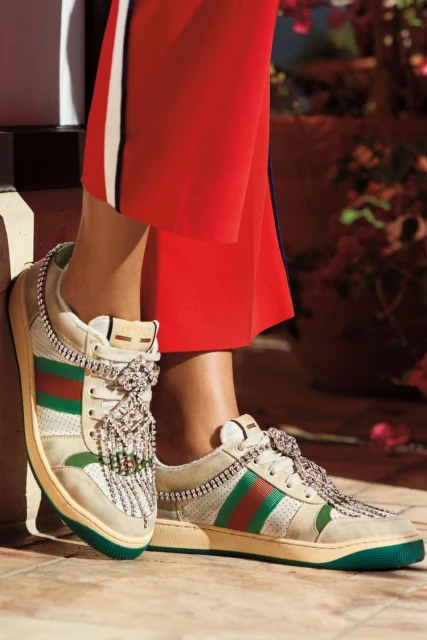 Gucci sneakers with jeweled laces, paired with red pants featuring a white stripe. The footwear is positioned on a tiled surface.