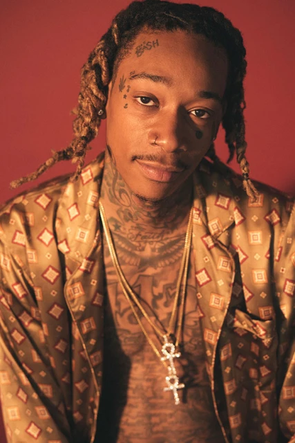 Close-up portrait of Wiz Khalifa, showcasing his blonde dreadlocks, tattoos, gold necklaces, and patterned shirt against a red backdrop.