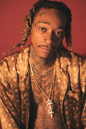 Close-up portrait of Wiz Khalifa, showcasing his blonde dreadlocks, tattoos, gold necklaces, and patterned shirt against a red backdrop.